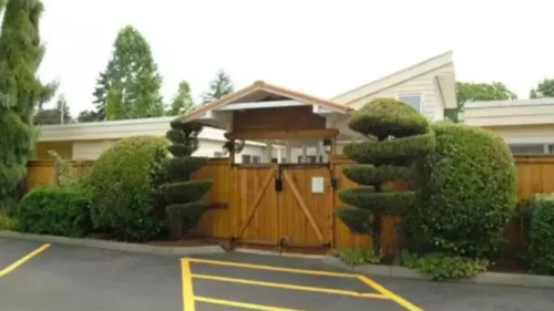 A wooden gate with a small covered roof, flanked by topiary bushes, leading into a building complex