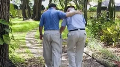 An elderly man using a cane is supported by a younger man with his arm around his shoulder as they walk down a path lined with trees